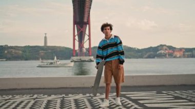 Young man posing skateboard at water view quay portrait. Thoughtful skater guy looking camera at Tagus river background closeup. Sporty hipster thinking about extreme trick at famous Lisbon place 