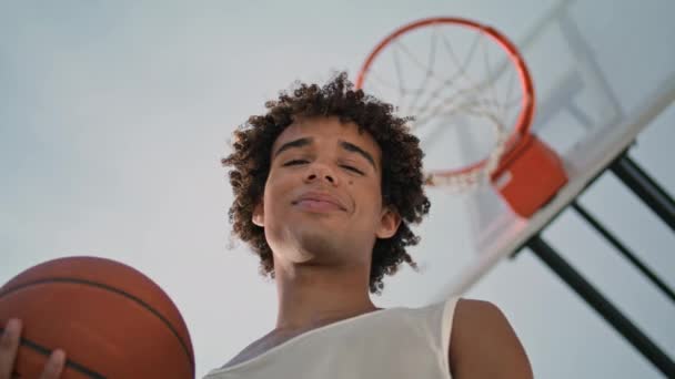 微笑的运动员在体育场的低角度持球 年轻的篮球运动员独自享受新鲜空气特写 阳光灿烂的早晨 卷曲的年轻人看着相机 快乐的少年在街上摆姿势 — 图库视频影像
