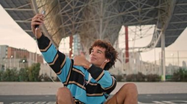 Positive blogger making selfie under bridge closeup. Smiling teen messaging smartphone at street. Curly guy listening earphones in happy mood outdoors. Joyful young man chatting social networks alone 