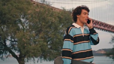 Friendly teenager speaking smartphone at Lisbon embankment. Curly guy talking mobile at evening bridge background. Contemporary skater walking at river view outdoor. Urban student ending call closeup