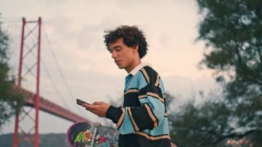 Smiling student walking mobile at coast closeup. Skater listening smartphone voice message outside. Curly hair zoomer putting phone to ear at river bridge place. Contemporary guy enjoying Lisbon