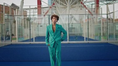 Curly guy smiling camera at modern stadium zoom on. Handsome young man in fashion suit looking attentively at urban background portrait. Well dressed model put hands in pockets closeup. Stylish person
