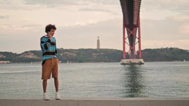 Positive hipster calling cellphone in Portugal city. Smiling guy using mobile on river embankment. Millennial student talking smartphone at urban background. Emotional teenager walking at famous place