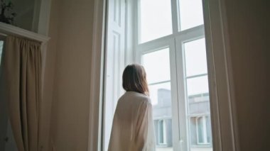 Young woman watching window at home morning closeup. Relaxed girl looking out street enjoying weekend back view. Dreaming lady in silk robe resting at light apartment. Domestic lifestyle concept