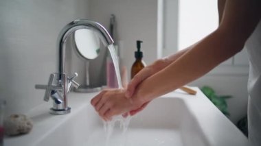 Girl washing hands at bathroom sink closeup. Unknown woman rubbing fingers under faucet at ceramics wash basin. Young lady cleaning arms alone at light modern apartment. Basic home hygiene concept