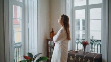 Thoughtful woman looking window at light interior. Relaxed girl watching street views enjoying weekend alone. Dreaming lady stretching hands resting at cozy apartment. Domestic lifestyle concept