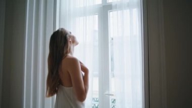 Sensual girl watching window at bedroom close up. Tender woman relaxing at white curtains place back view. Seductive young lady in nightgown touching her soft skin at cozy morning apartment alone