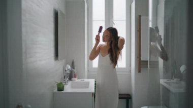 Calm girl holding hairbrush in bathroom alone. Confident woman doing morning hair care routine at modern apartment place. Gorgeous lady looking mirror standing at home. Domestic hairstyling concept