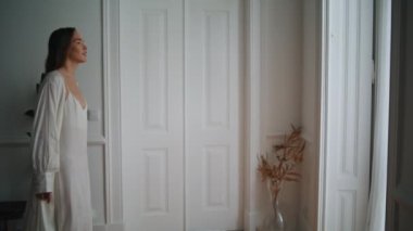 Relaxed woman looking window at morning indoors. Tender girl peeping from curtain at bedroom. Young lady in silk nightgown walking calmly at cozy apartment. Female person spending lazy weekend