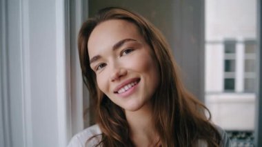 Cheerful woman smiling camera at home portrait. Positive girl playing hair at white window interior closeup. Sensual lady relaxing alone posing at morning apartment. Tender model resting at weekend