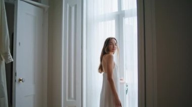 Tender lady posing window interior. Sensual woman looking camera relaxing at white curtains place. Longhaired girl enjoying calm morning at cozy bedroom. Female person standing in dark apartment alone