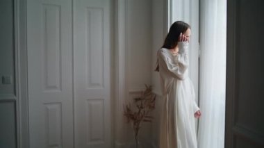 Young woman watching window at home morning. Relaxed model looking out street enjoying calm weekend. Dreaming lady in silk robe resting alone at light apartment interior. Domestic lifestyle concept