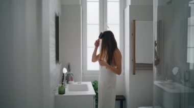 Attractive model combing hair at mirror place. Confident woman doing hairstyle in modern bathroom alone. Gorgeous girl getting ready making everyday morning routine at home. Daily beauty procedures