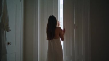 Tender lady opening shutters at bedroom. Relaxed woman looking window admiring views at home weekend. Unknown girl in silk nightgown enjoying lazy morning alone at dark place interior. New day concept