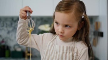 Focused child holding whisk at kitchen closeup. Little girl mixing ingredients making dough. Cute baby in apron combining carefully in bowl. Junior chef kneading attentively preparing baking alone