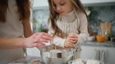 Focused baby mixing whisk at kitchen closeup. Smiling mother holding dish helping to daughter. Cute child in apron combining ingredients in bowl. Positive family cooking together at light interior