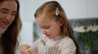 Young mother teaching daughter cooking indoor portrait. Focused baby in apron making dough at kitchen closeup. Loving woman helping to kid bake at home. Positive family kneading together at cook room