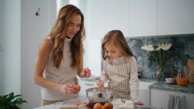 Smiling mother daughter breaks eggs at kitchen. Adorable child mixing ingredients at bowl helping mom at light place. Positive family cooking together at home interior. Lady and baby in domestic place