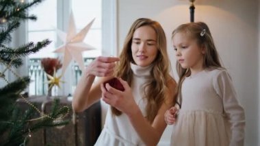 Sevgili anne kızı Noel ağacını kapalı mekanda dekore ediyor. Gülümseyen kadın rahat bir odada bebeğe Xmas oyuncağı veriyor. Yeşil köknar dalına süsleme yapan meraklı küçük kız. Yeni yıl konsepti