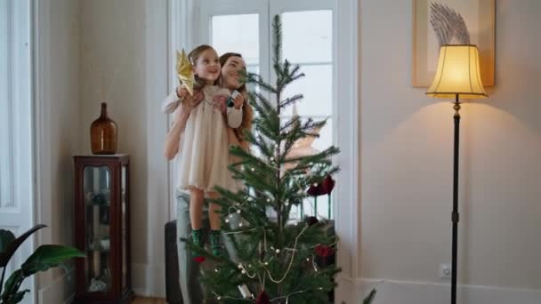可敬的家人正在准备圣诞节的家庭特写 笑着妈妈接女儿用柔情放大 好奇的小女孩把圣诞树放在客厅的树梢上 新年筹备概念 — 图库视频影像