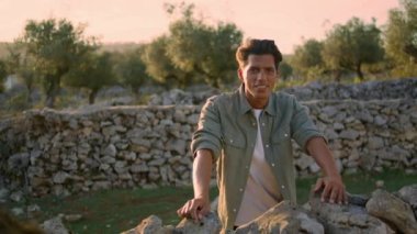 Rahatlamış bir adam, doğa portresine taş duvara yaslanıyor. Yazlık mekanda gülümseyen, bronzlaşmış bir adam kameraya yakından bakıyor. Sadece zeytin ağaçları çiftliğinde poz veren pozitif işçi. Halk konsepti