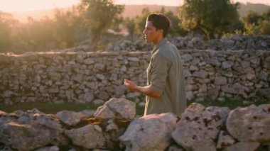 Düşünceli adam taş duvarda yürüyormuş. Yakından. Ciddi bir çiftçi tek başına karşıya geçiyor güneş ışınları kırsal alanda dinleniyor. Odaklanmış adam zeytin ağaçlarının eski çit manzarasını izliyor.
