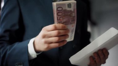 Unknown man putting money cash into white paper envelope close up. Elegant businessman wearing suit placing pack of euro bills in envelope indoors. Business payments financial profit wealth concept.