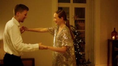 Carefree pair dancing at New Year celebration home closeup. Positive couple holding hands moving together at Christmas tree interior. Smiling man twirling woman in party dress at night apartment