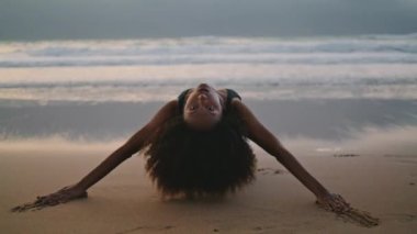 Sexy woman dancer performing sensual movements on beach lying sand at twilight. Attractive flexible african american girl in black swimsuit bending body seductively. Curly lady dancing in darkness.