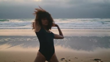 Seductive african american girl dancing sensual dance on wet beach sand cloudy evening. Curly flexible woman performing contemporary dance on seashore summer twilight. Attractive brunette moving body.