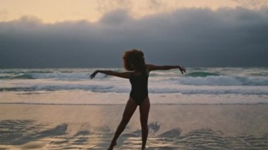 African american attractive girl dancing on beach at evening darkness wearing black swimsuit. Talented woman performer moving body gracefully on wet sand. Curly brunette performing modern style.