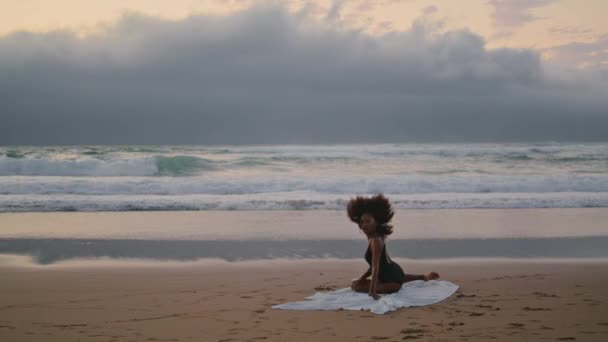 陰鬱なビーチで砂に座ってダンスを行う精力的な魅力的な女性 海の波の近くの海岸で優雅に踊るアフリカ系アメリカ人の少女 ブルネットは優雅にフロントグレーの空に移動 — ストック動画