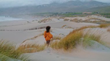 Aktif, çıplak ayaklı bir kız bulutlu bir akşamda kum tepelerinde kuru çimlerle koşuyor. Kıvırcık Afrika kökenli Amerikalı bir kadının boş deniz kıyısında koştuğunu parlak turuncu elbiseler giydiğini kasvetli bir havada.