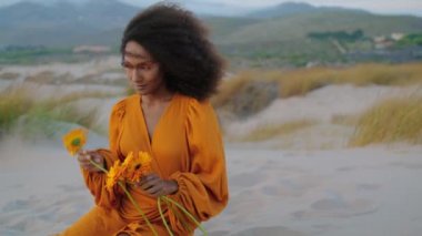 Alacakaranlıkta, kumlu çölde portakal çiçeği koklayan Afro-Amerikan modelinin portresi. Huzurlu gülümseyen kameralı rüya gibi çekici kadın. Şık elbiseli kıvırcık esmer, kumullarda oturuyor..