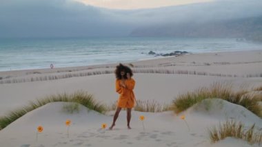 Genç, esnek bir kadın kumsalda, turuncu çiçeklerin yanında, kasvetli bir akşamda zarifçe hareket ediyor. Kıvırcık Afro-Amerikan kızı çağdaş tarzda dans ediyor Alacakaranlıkta deniz kıyısında parlak elbiseler giyiyor. 