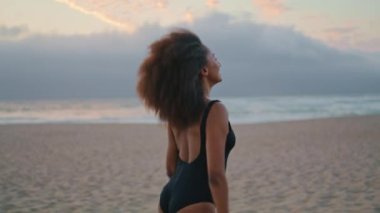 Relaxed woman tourist walking sandy gloomy beach in black swimsuit at summer dusk. Carefree african american girl relaxing on beautiful seashore at twilight. Attractive young brunette going near sea.
