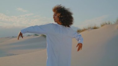 Yaz rüzgarlı akşamlarda kumlu çölde modern dans eden odaklanmış genç bir kadın. Sakin Afrikalı Amerikalı kız beyaz elbise giyerek anlamlı hareketler yapıyor. Doğadaki çağdaş performans.