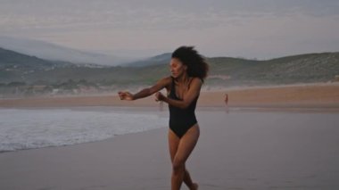 Sensual african american woman dancing modern style on overcast beach wearing black swimsuit. Slim curly girl making smooth hands movements walking on wet sand. Sexy brunette performing at gloomy sky.