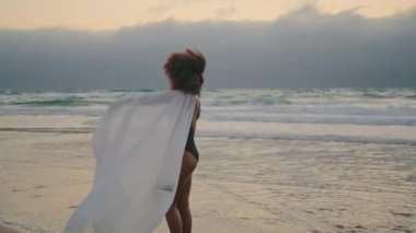 Sad african american woman on overcast beach letting go white shawl. Calm curly girl wearing black swimsuit standing on sand seashore holding satin pareo. Young brunette feeling lonely at cloudy day.