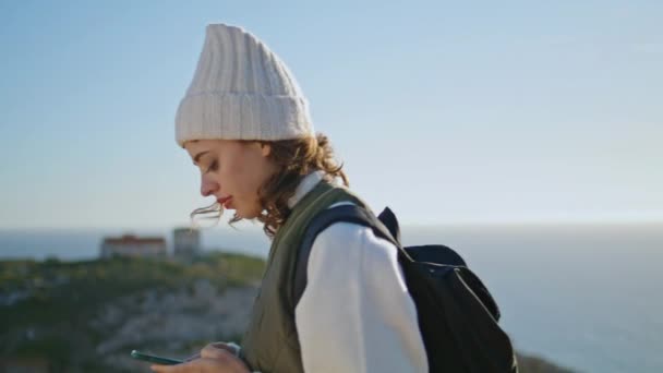 海の山の景色でスマートフォンをテキストメッセージハイキング女の子 笑顔の観光客のバックパックと一緒に旅行歩く 旅行中のウェブ検索ナビゲーションを閲覧するハイカー女性に焦点を当てました オンラインコミュニケーションの概念 — ストック動画