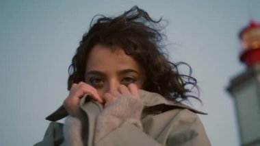 Dreamy girl rest seaside lighthouse view closeup. Happy romantic woman get warm hiding adjusting coat on cold windy evening. Beautiful smiling tourist look camera enjoy night. Stress free calm female