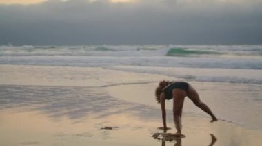 Energetic girl dancer moving flexible body on wet sand beach wearing black swimsuit at gloomy evening. Passionate curly african american brunette making acrobatic wheel performing dance on seashore.