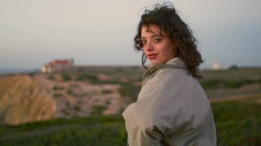 Smiling woman posing ocean evening cliff. Carefree lonely tourist enjoy calm landscape looking camera. Beautiful curly girl taking break relaxing seaside on vacation. Stress free lifestyle concept.