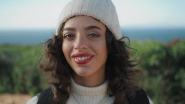 Cheerful girl looking camera on windy day closeup. Happy young traveler resting ocean shore enjoy spring vacation. Smiling pretty woman tourist posing feel joyful in knitted hat. Outdoors trip leisure