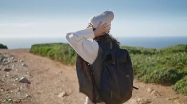 Tourist walking rocky pathway at ocean shore. Relaxed girl going with backpack admiring spring nature. Young curly woman exploring traveling seaside on windy day. Contemplating life solitude concept.