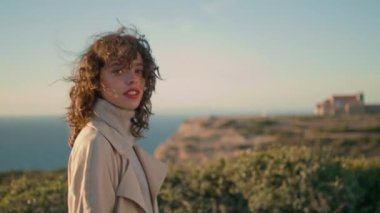 Casual woman going evening ocean mountain. Serene girl looking camera enjoying calm weekend. Cheerful millennial touching curly hair spinning at beautiful sea view. Femininity natural beauty concept.