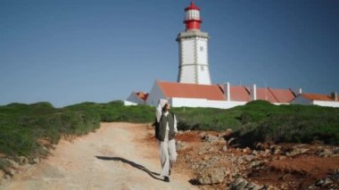 Aktif kız güneşli bir günde deniz feneri manzarasında yürüyor. Deniz kenarı gezisinde dinlenirken mutlu turistler. Sırt çantasıyla gezintiye çıkan güzel bir kız öğrenci. Tasasız yalnız gezgin manzaraya hayrandır.