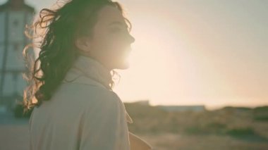 Closeup casual girl walking in sun. Stylish beautiful woman enjoying evening on windy seashore island. Curly female silhouette dreaming looking camera at lighthouse view. Serene satisfied tourist.