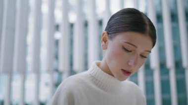 Portrait of thoughtful attractive girl looking down in front modern building outdoors. Young pensive woman reading typing messaging alone on street close up. Beautiful lady face with elegant hairstyle