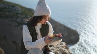 Calm girl enjoying book at ocean cliff edge. Focused traveler resting mountain reading literature on windy spring day. Romantic woman reader relax outdoors spend weekend at sea. Hobby travel concept